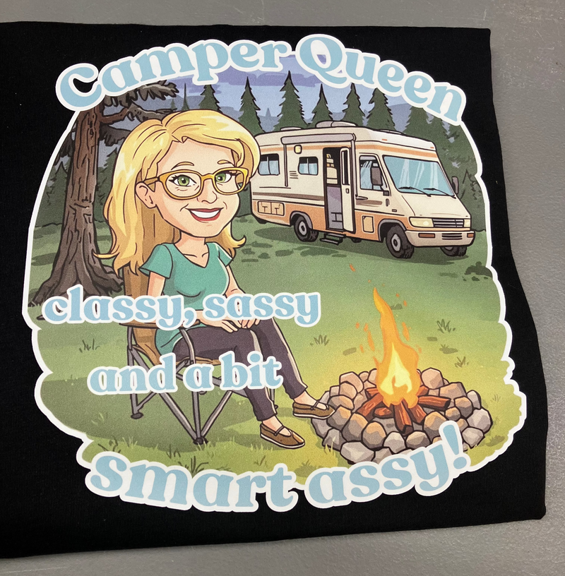 Black t-shirt with a graphic of a woman by a campfire and an RV, text 'Camper Queen classy, sassy and a bit smart assy!' on a gray background.
