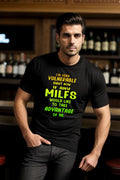 Black "Searching for a MILF" Tee with colorful text, worn by a person in a bar setting.