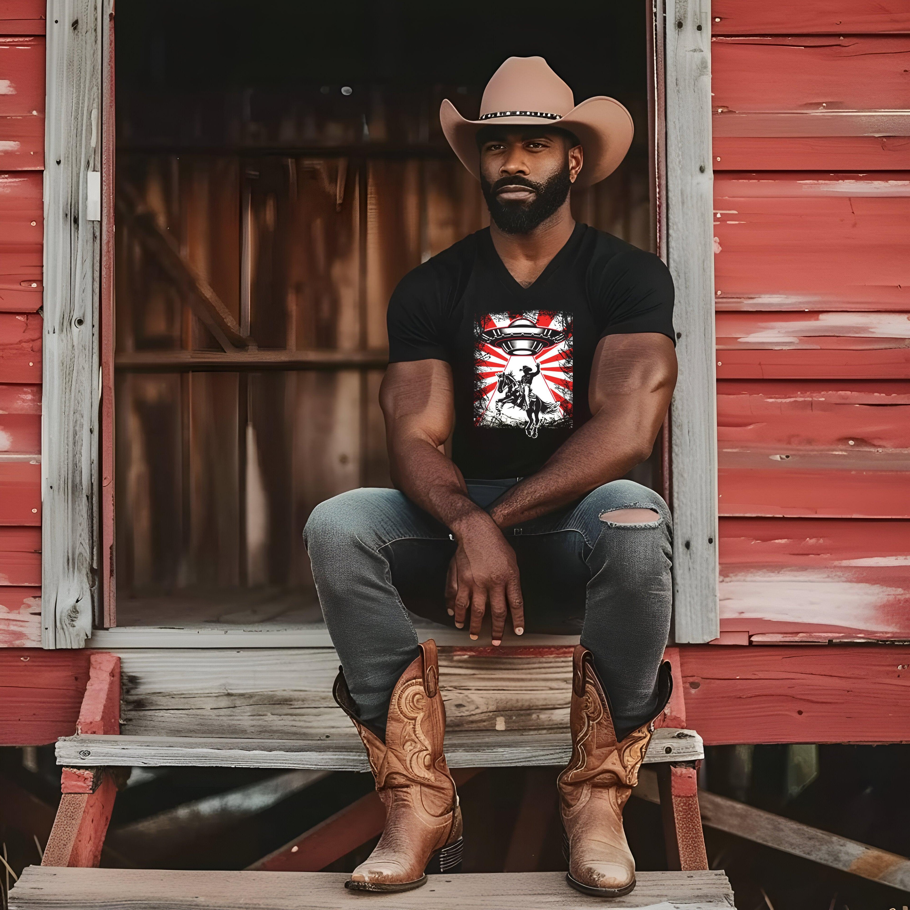 UFO Cowboy Abduction Unisex Tee with graphic design worn by man sitting on wooden steps, cowboy hat and boots.